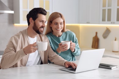 Photo of Happy couple with laptop and cups of drink at white table in kitchen