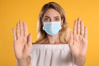 Photo of Woman in protective face mask showing stop gesture on yellow background. Prevent spreading of coronavirus