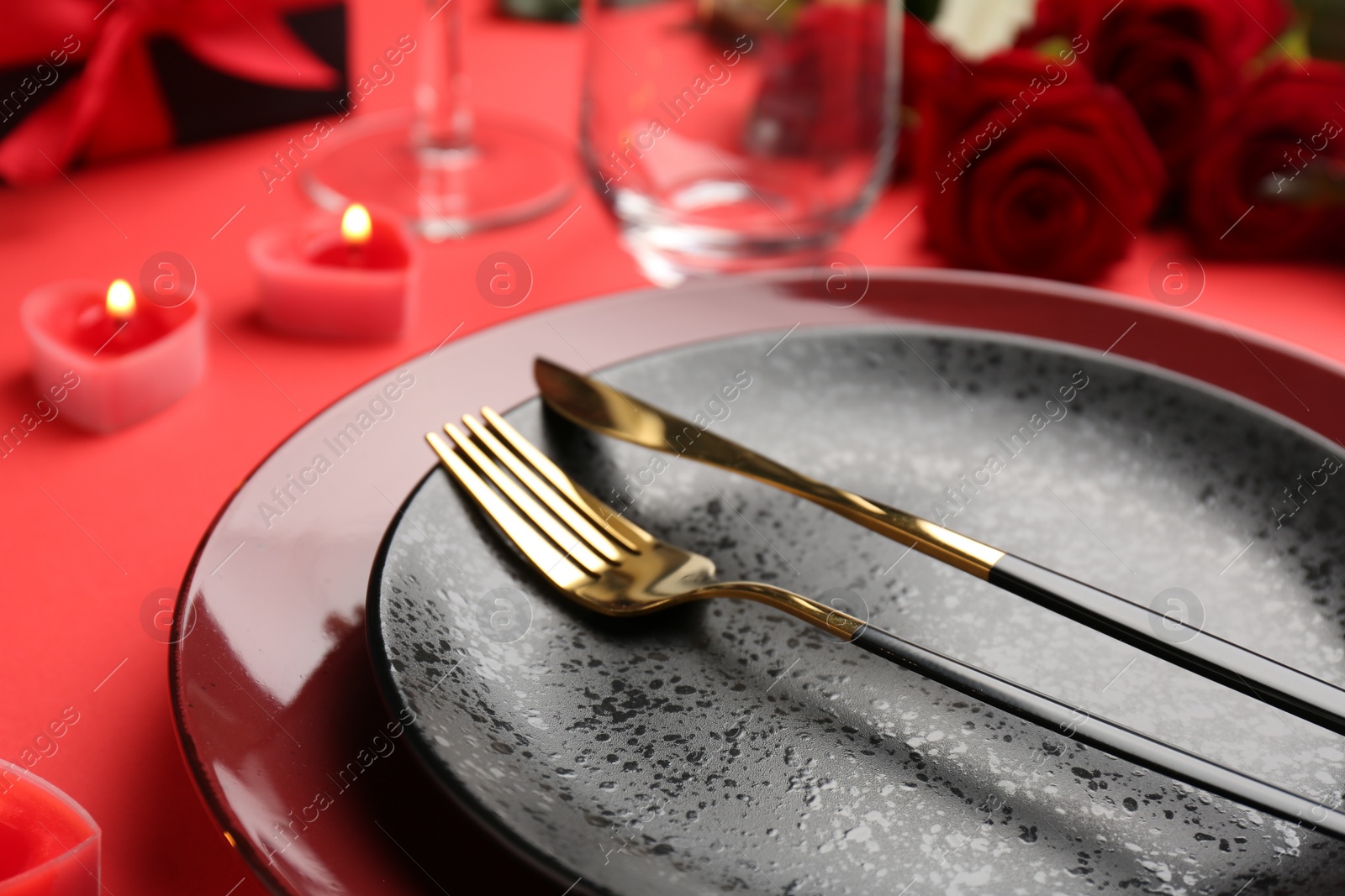 Photo of Place setting with heart shaped candles, gift box and bouquet of roses on red table, closeup. Romantic dinner