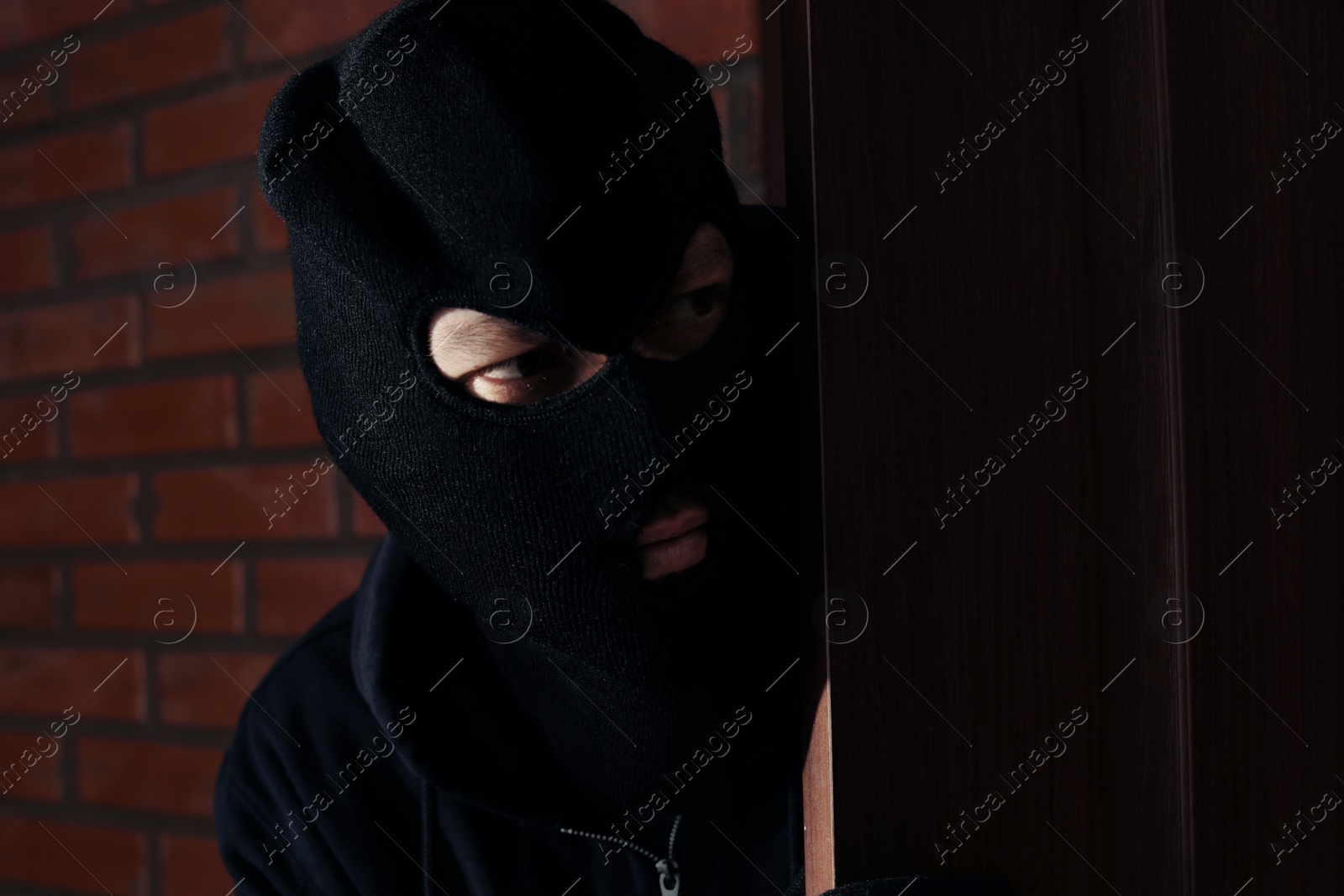 Photo of Man in mask spying behind door indoors. Criminal activity