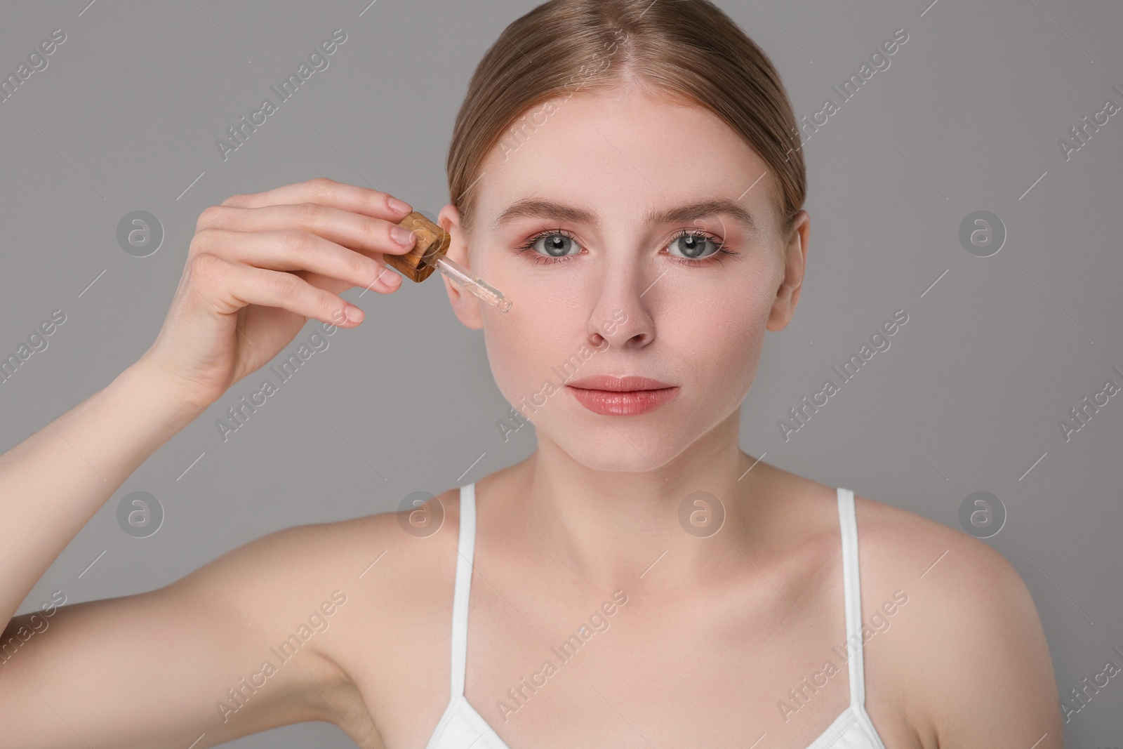 Photo of Beautiful young woman applying essential oil onto face on grey background