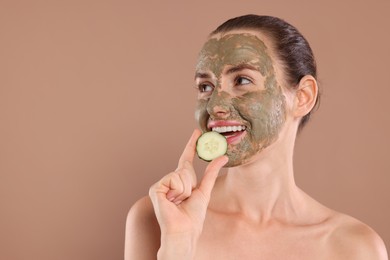 Photo of Beautiful woman with clay mask holding piece of cucumber on light brown background, space for text