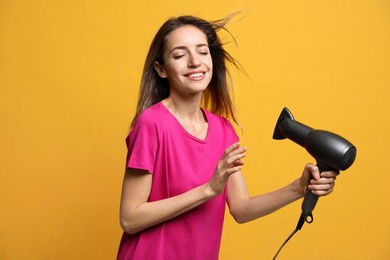 Beautiful young woman using hair dryer on yellow background