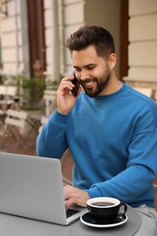 Handsome young man talking on smartphone while using laptop at table in outdoor cafe