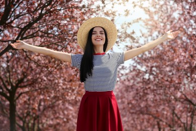 Photo of Pretty young woman with straw hat near beautiful blossoming trees outdoors. Stylish spring look