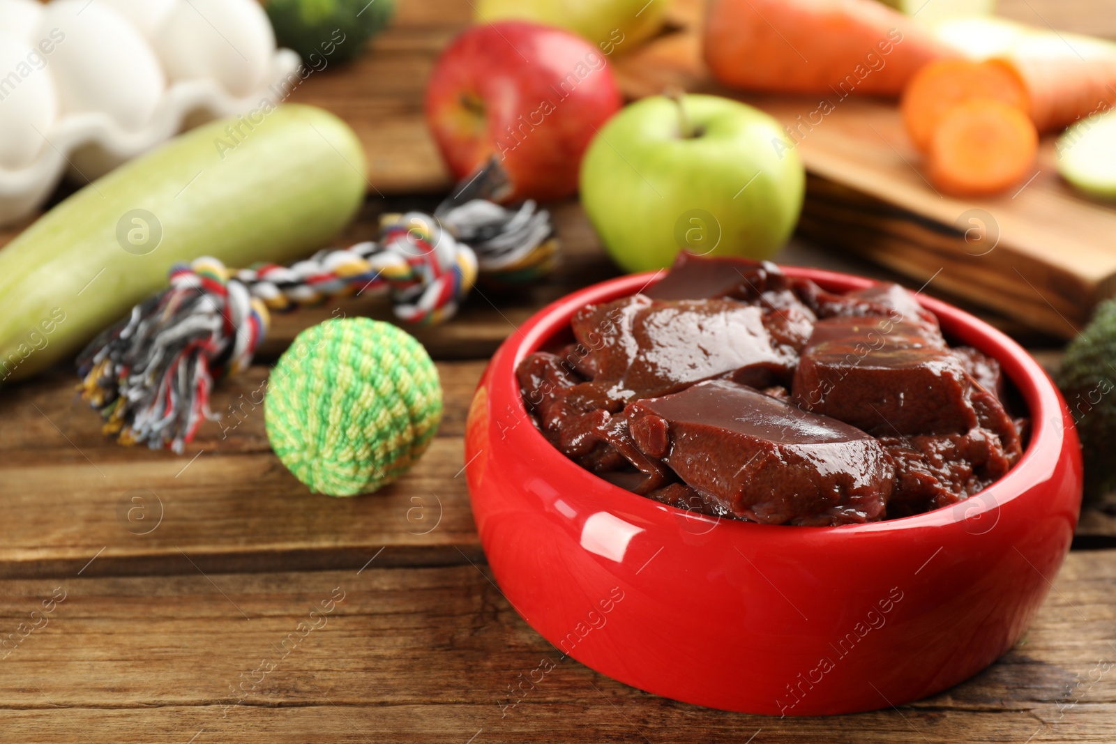 Photo of Feeding bowl with liver and toys on wooden table, closeup. Natural pet food