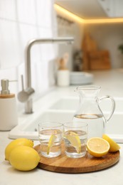 Jug, glasses with clear water and lemons on white table in kitchen