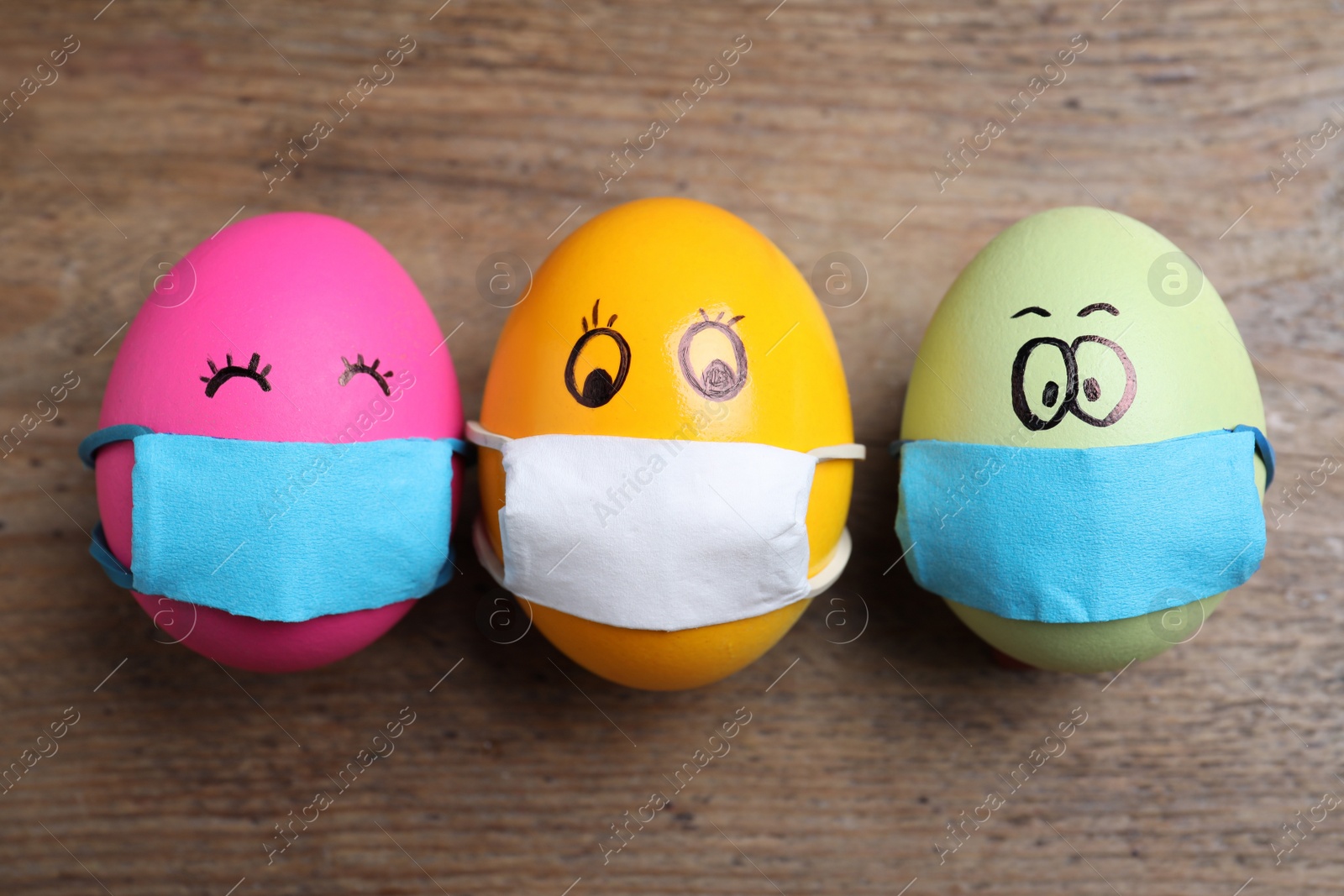 Photo of Bright eggs in protective masks on wooden table, flat lay. Easter holiday during coronavirus quarantine