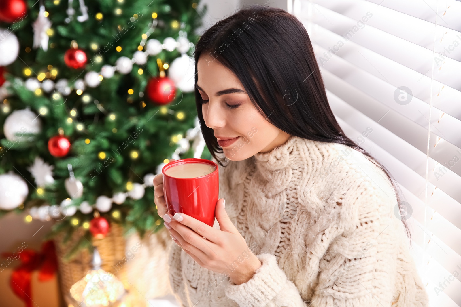 Photo of Young woman holding cup of coffee in room with Christmas tree