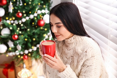 Photo of Young woman holding cup of coffee in room with Christmas tree