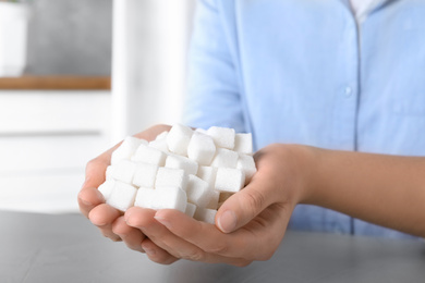 Woman holding heap of refined sugar cubes at table indoors, closeup