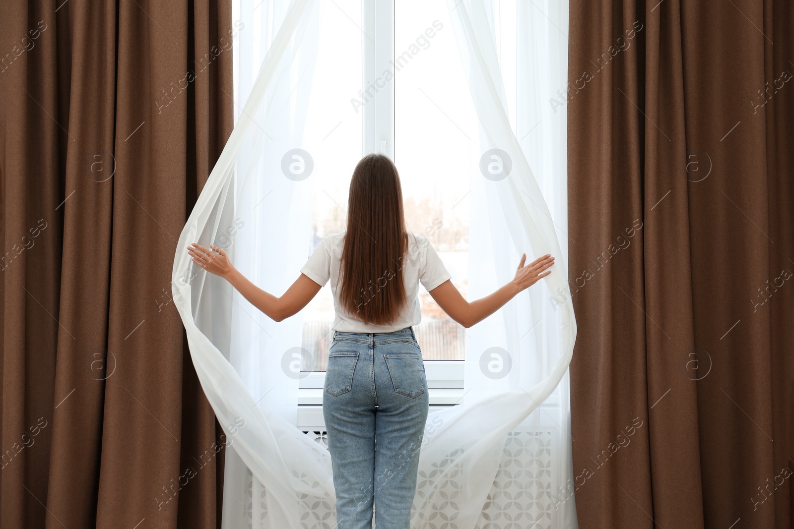 Photo of Woman opening elegant window curtains in room, back view