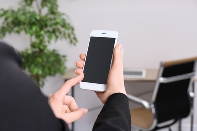 Woman holding mobile phone with blank screen in office, closeup. Space for text