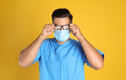 Doctor wiping foggy glasses caused by wearing medical mask on yellow background