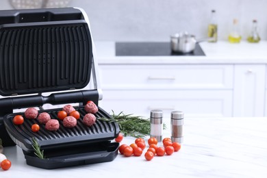 Photo of Electric grill with meatballs, tomatoes, spices and vegetables on white marble table in kitchen. Space for text