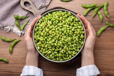 Woman holding bowl of edamame beans at wooden table, top view