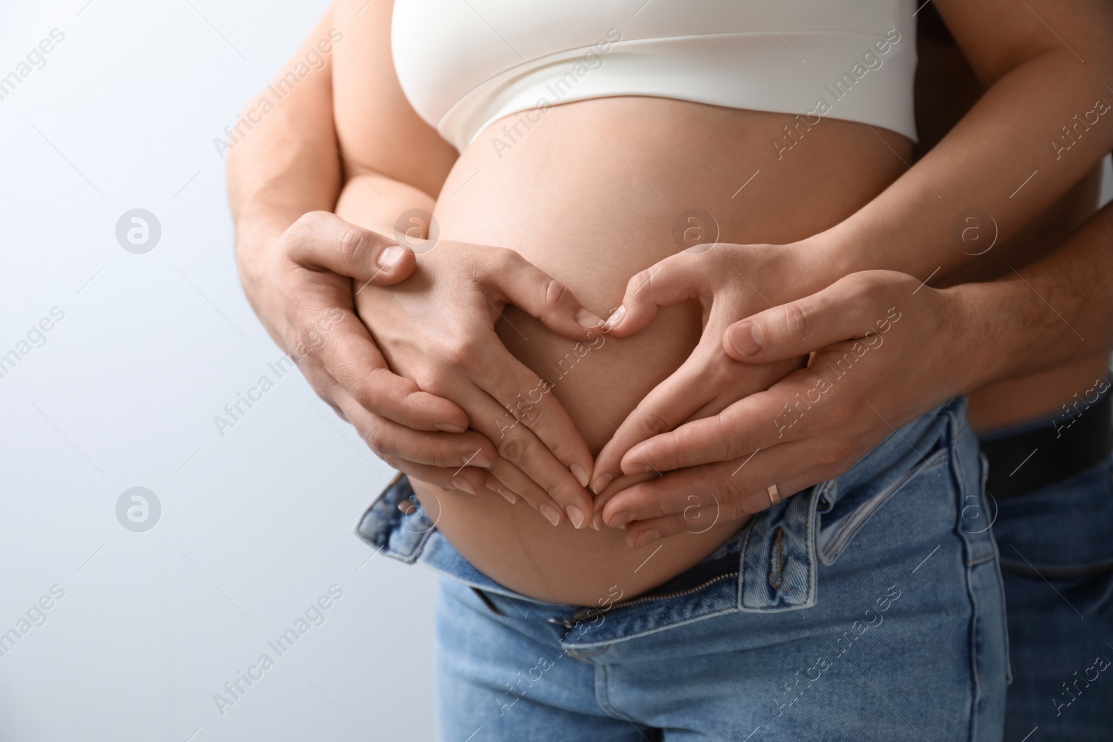 Photo of Pregnant young woman making heart with hands on belly and husband near her against light background, closeup