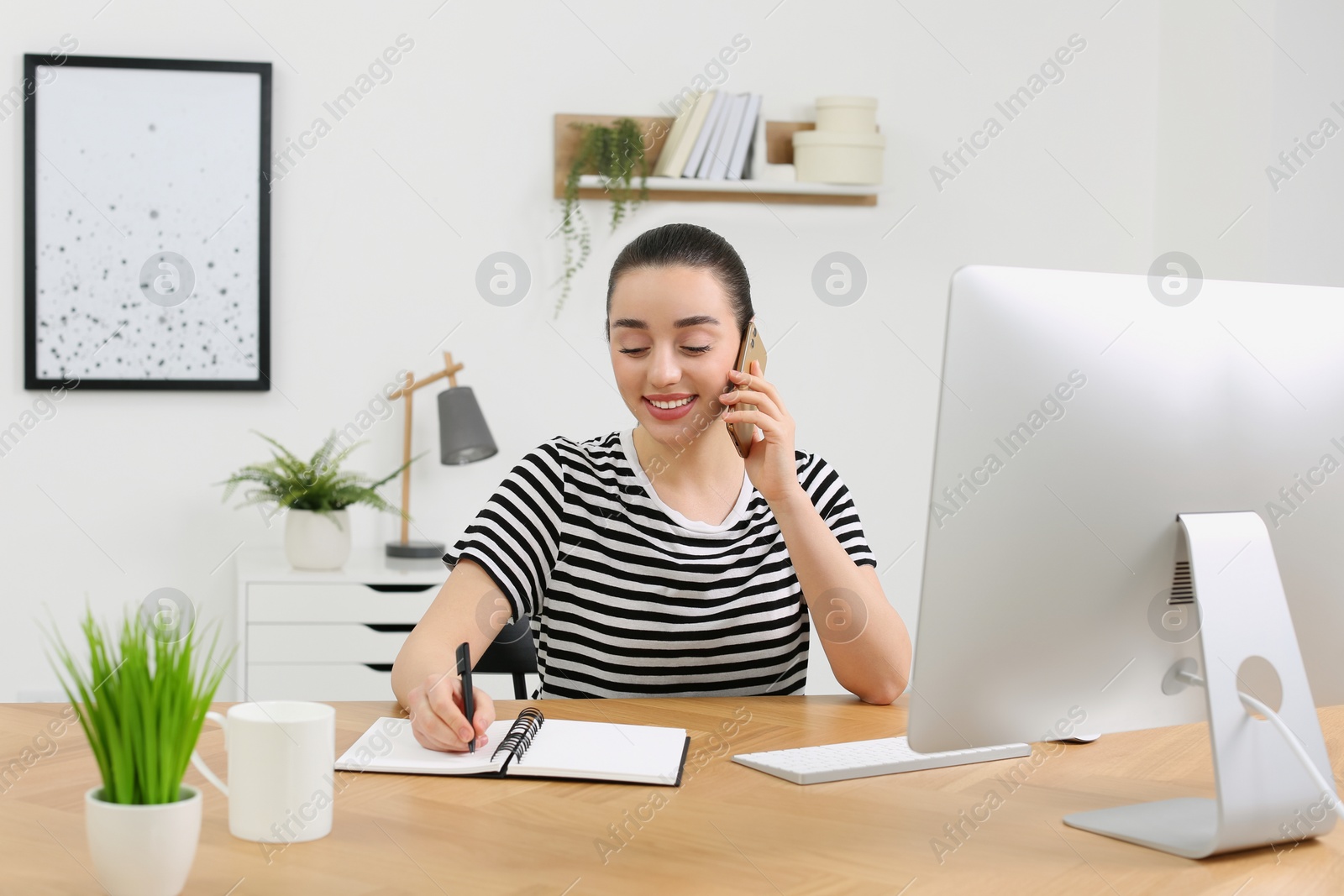 Photo of Home workplace. Happy woman with pen and notebook talking on smartphone at wooden desk in room