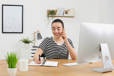 Photo of Home workplace. Happy woman with pen and notebook talking on smartphone at wooden desk in room