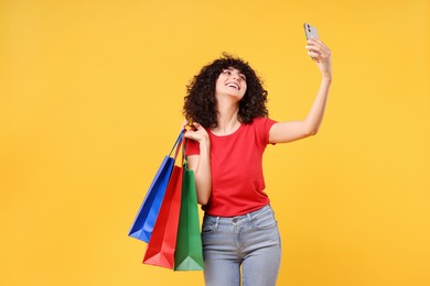 Happy young woman with shopping bags taking selfie on yellow background