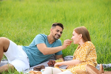 Photo of Young man feeding his girlfriend with grape on picnic blanket outdoors