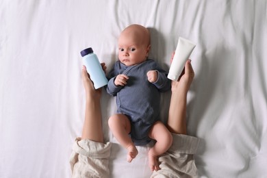 Mother holding body cream and dusting powder near cute little baby on bed, top view