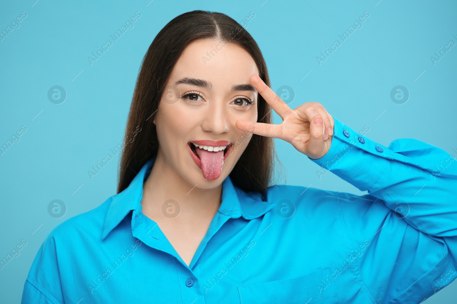 Photo of Happy woman showing her tongue and V-sign on light blue background