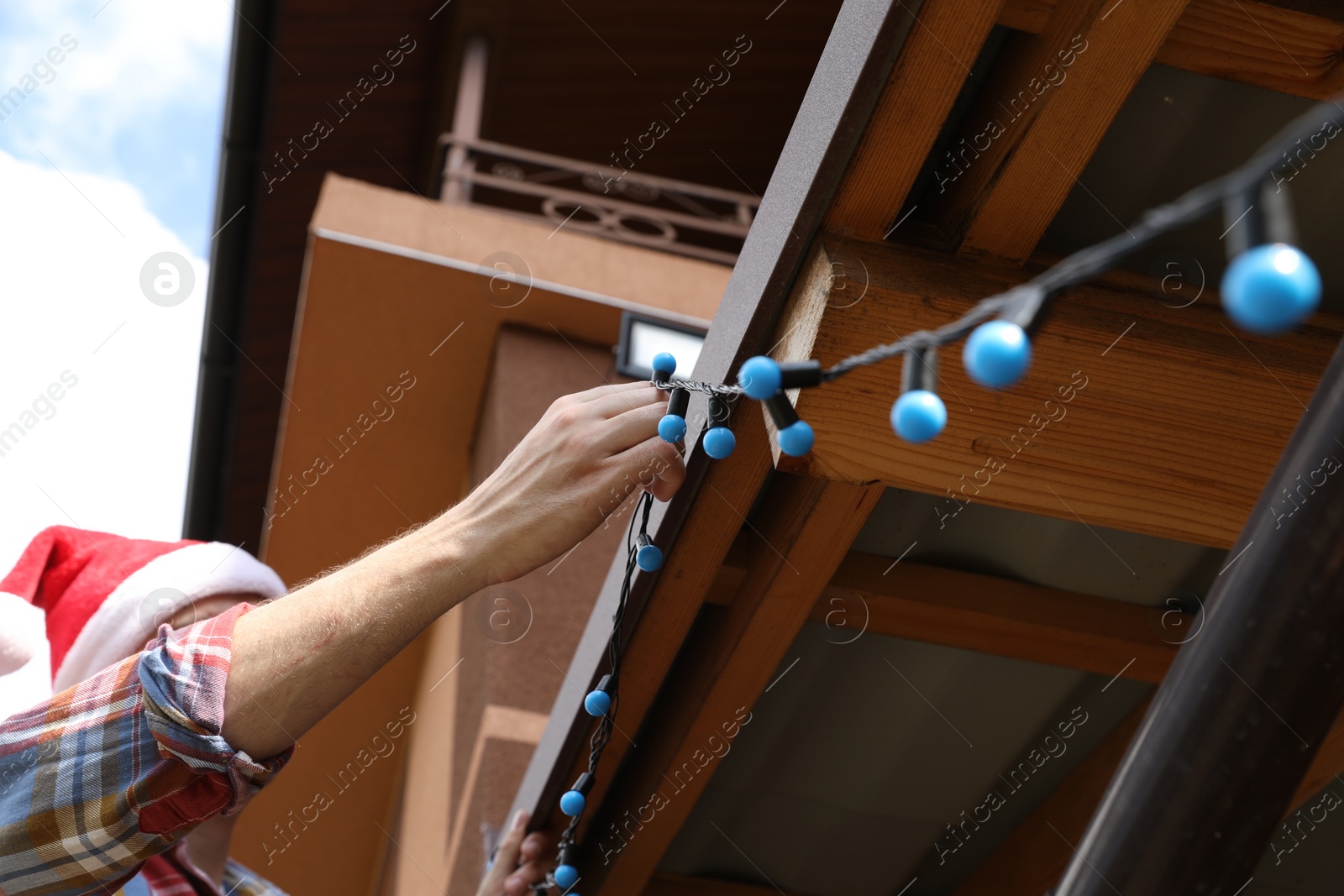 Photo of Man in Santa hat decorating house with Christmas lights outdoors, low angle view