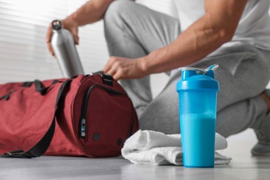 Photo of Young man putting bottle of water into bag indoors, focus on protein shake and towel