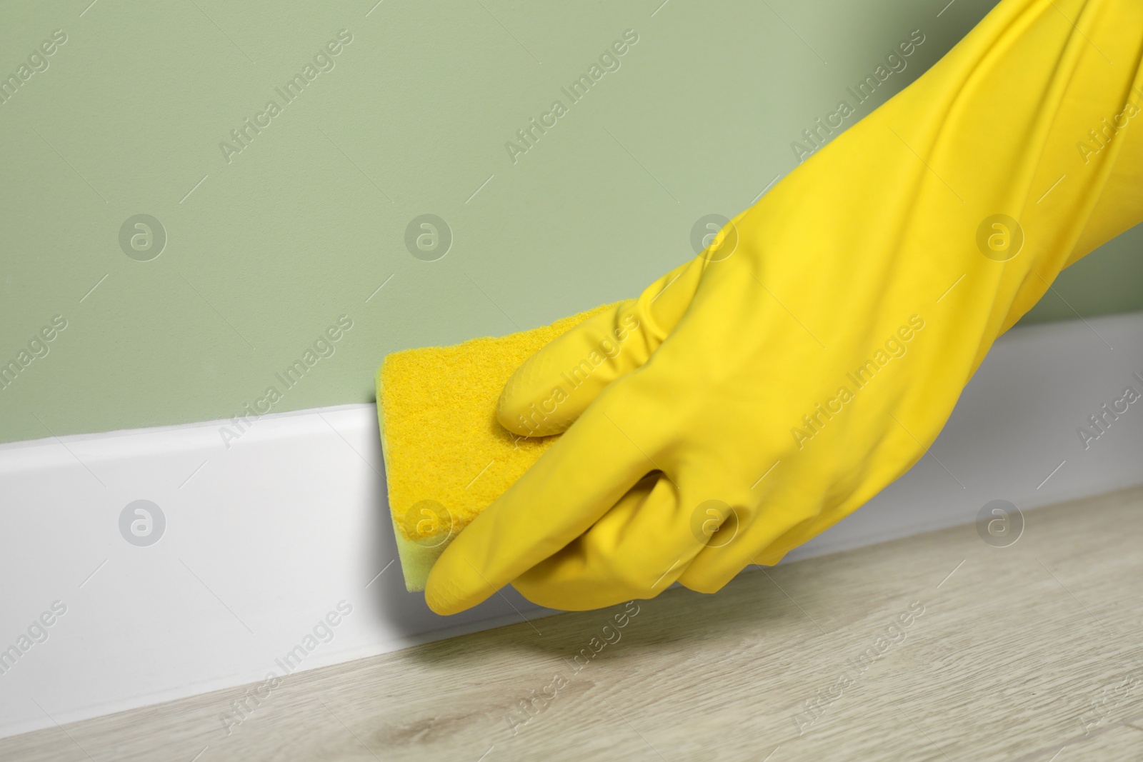 Photo of Woman in protective glove cleaning plinth with sponge indoors, closeup