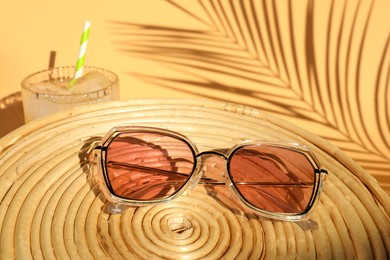 Photo of Stylish sunglasses on wicker mat near glass of refreshing drink against beige background