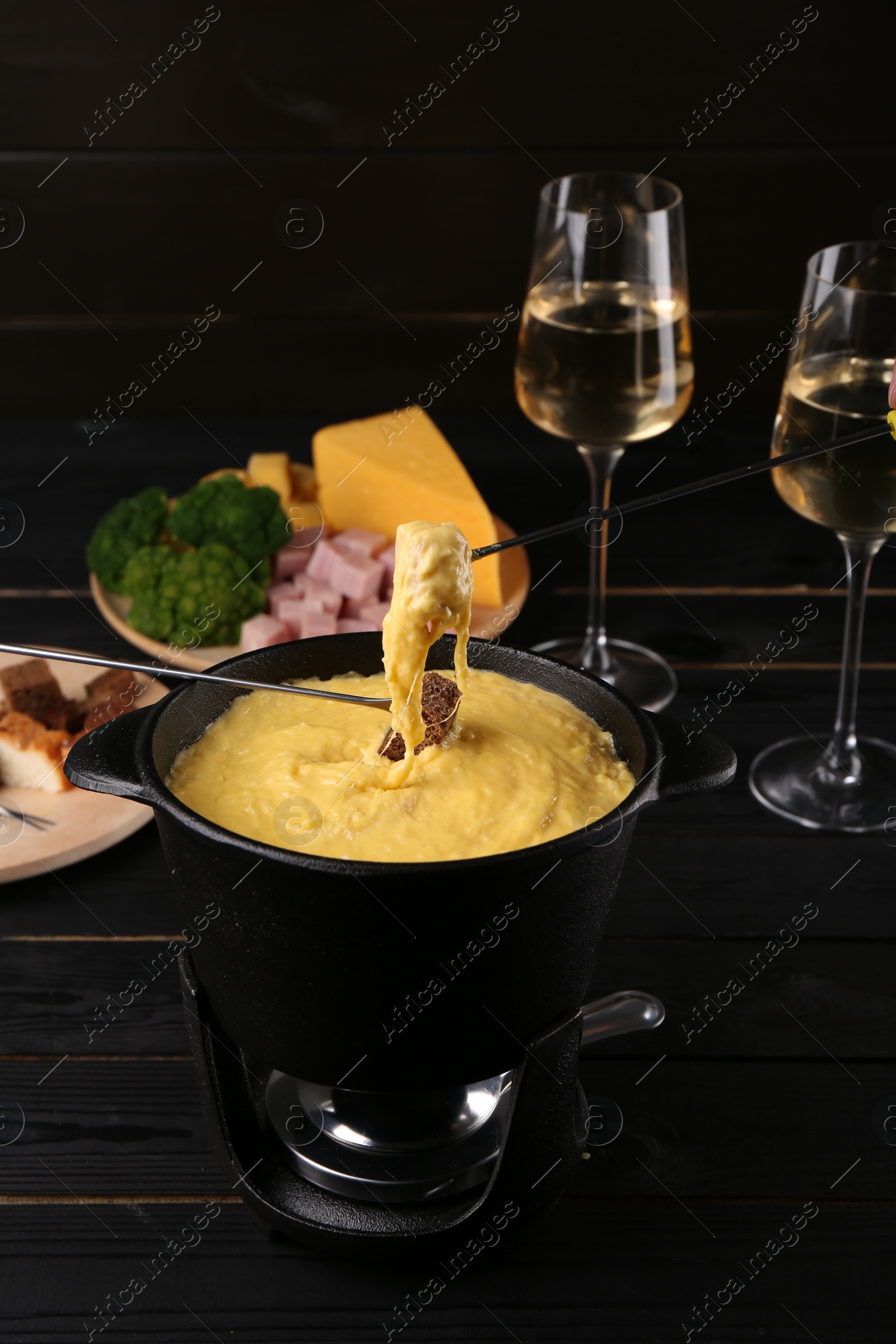 Photo of Dipping pieces of ham and bread into fondue pot with melted cheese on black wooden table