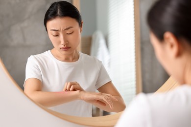 Photo of Suffering from allergy. Young woman scratching her arm near mirror in bathroom