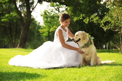 Bride and adorable Golden Retriever wearing wreath made of beautiful flowers on green grass outdoors