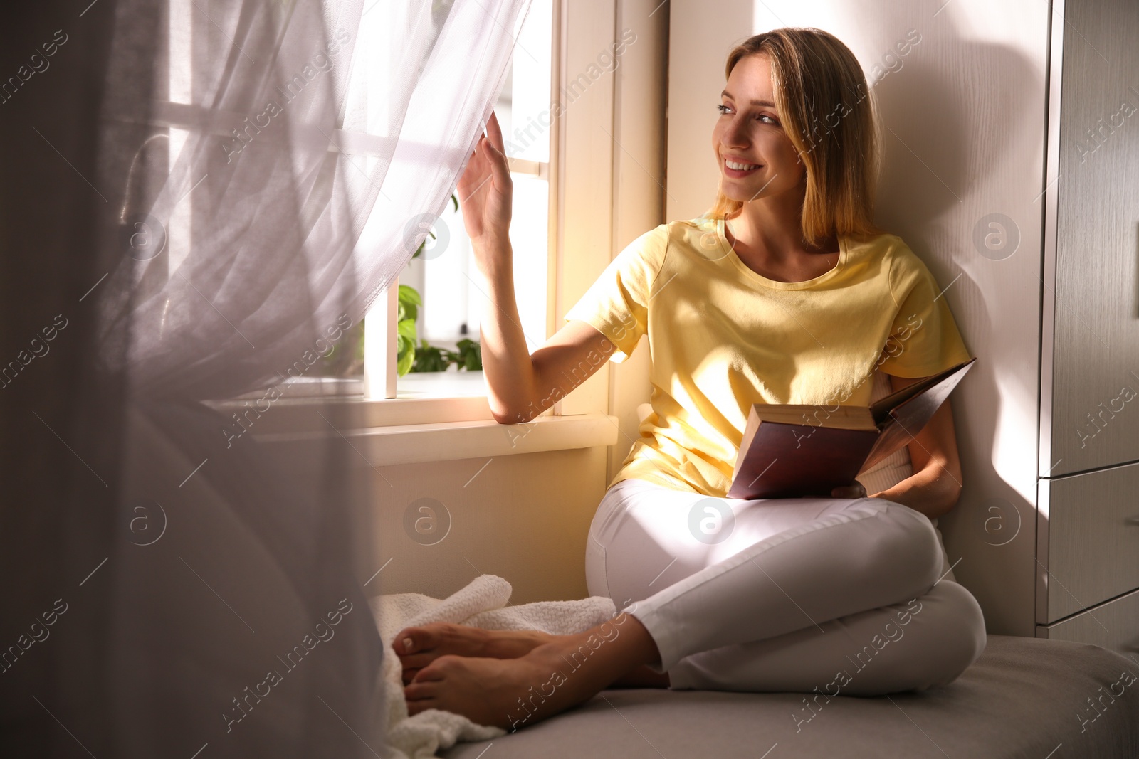 Photo of Beautiful young woman reading book near window at home