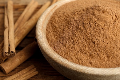 Photo of Aromatic cinnamon powder in bowl and sticks on wooden table, closeup