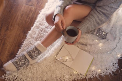 Woman with cup of hot beverage and book at home in winter evening, closeup