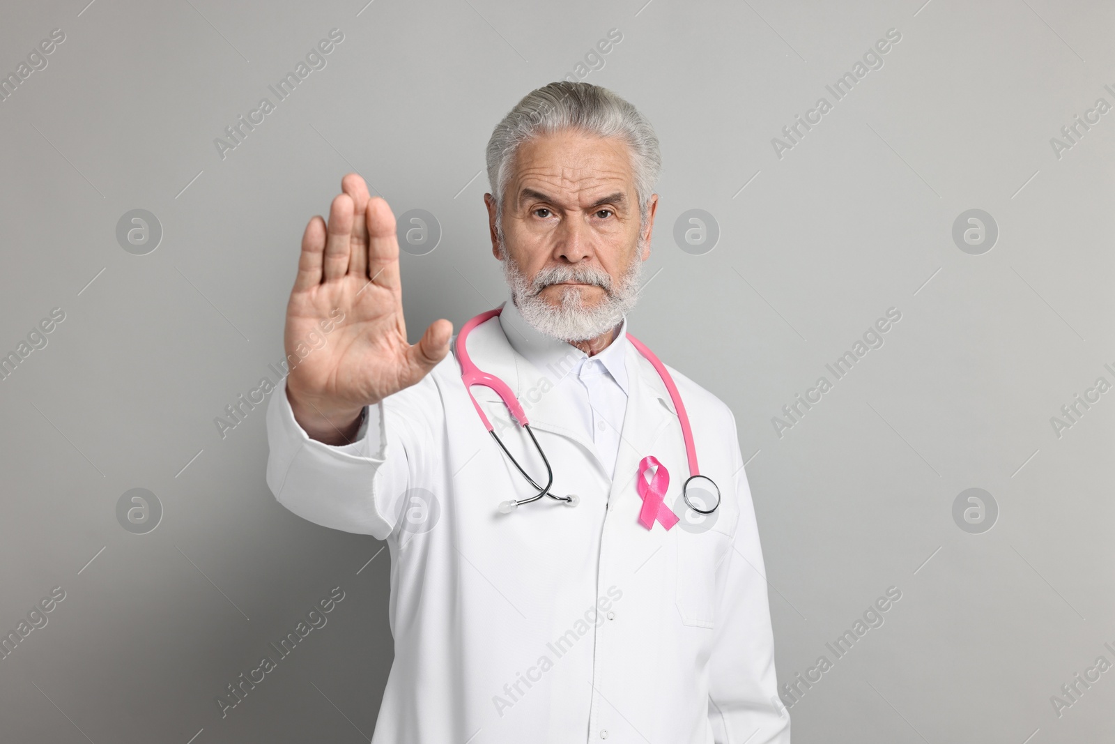 Photo of Mammologist with pink ribbon showing stop gesture on gray background. Breast cancer awareness