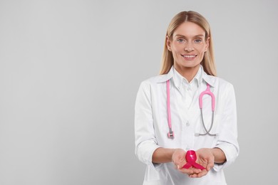 Photo of Doctor with stethoscope holding pink ribbon on light grey background, space for text. Breast cancer awareness