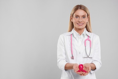 Doctor with stethoscope holding pink ribbon on light grey background, space for text. Breast cancer awareness