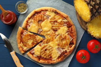 Delicious cut pineapple pizza, ingredients and knife on blue wooden table, flat lay