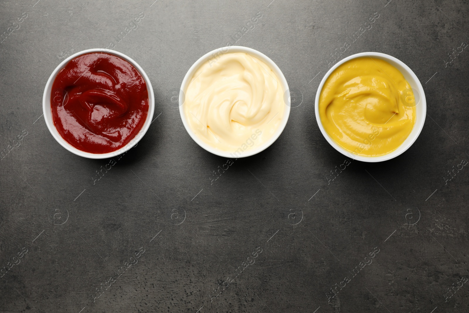 Photo of Ketchup, mustard and mayonnaise in bowls on grey table, flat lay. Space for text