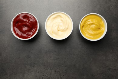 Photo of Ketchup, mustard and mayonnaise in bowls on grey table, flat lay. Space for text