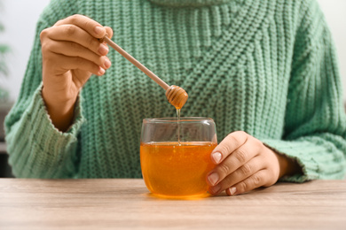 Photo of Woman holding honey dipper over jar at wooden table, closeup