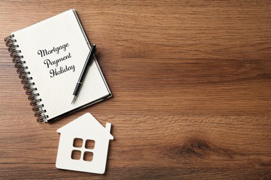 Photo of Notebook with words Mortgage Payment Holiday, pen and house model on wooden table, flat lay. Space for text