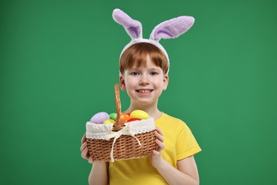 Photo of Easter celebration. Cute little boy with bunny ears and wicker basket full of painted eggs on green background