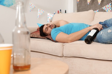 Young man with bottle of wine sleeping on sofa in messy room after party