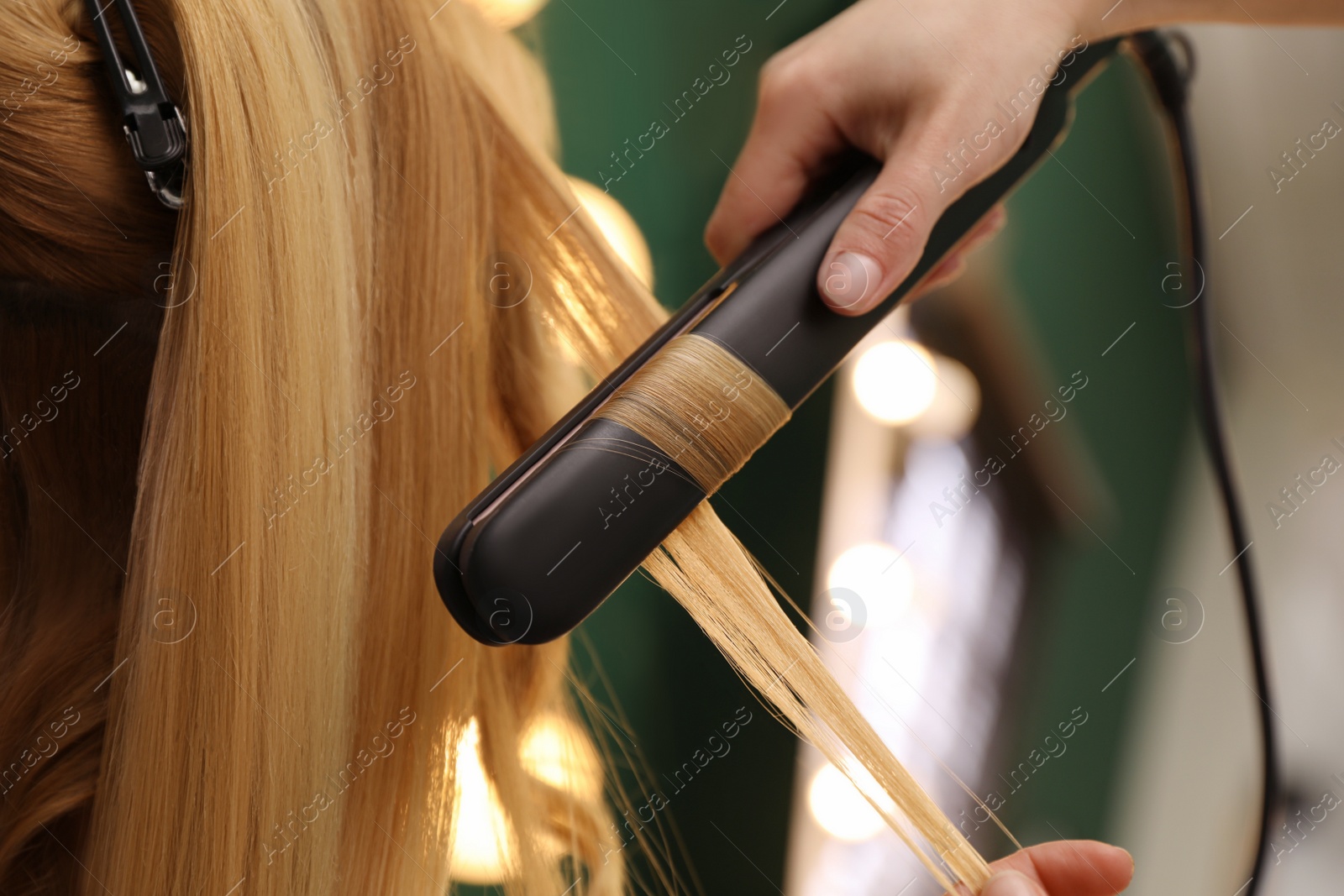Photo of Stylist curling woman's hair with flat iron in salon, closeup