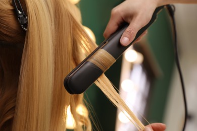 Photo of Stylist curling woman's hair with flat iron in salon, closeup