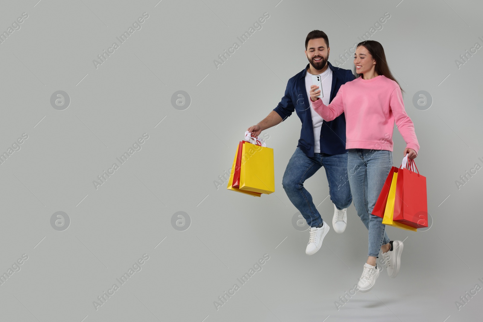 Photo of Happy couple with shopping bags and smartphone jumping on grey background. Space for text